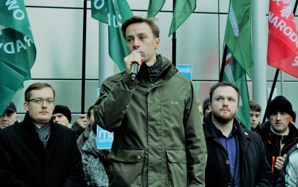 Foto von Krzysztof Bosak als Redner bei einer Veranstaltung der neofaschistischen Organisation Ruch Narodowy und der von Facebook gebannten neofaschistischen Organisationen ONR und MW. Im Hintergrund hinter Bosak sind die entsprechenden Flaggen von Ruch Narodowy und ONR erkennbar.